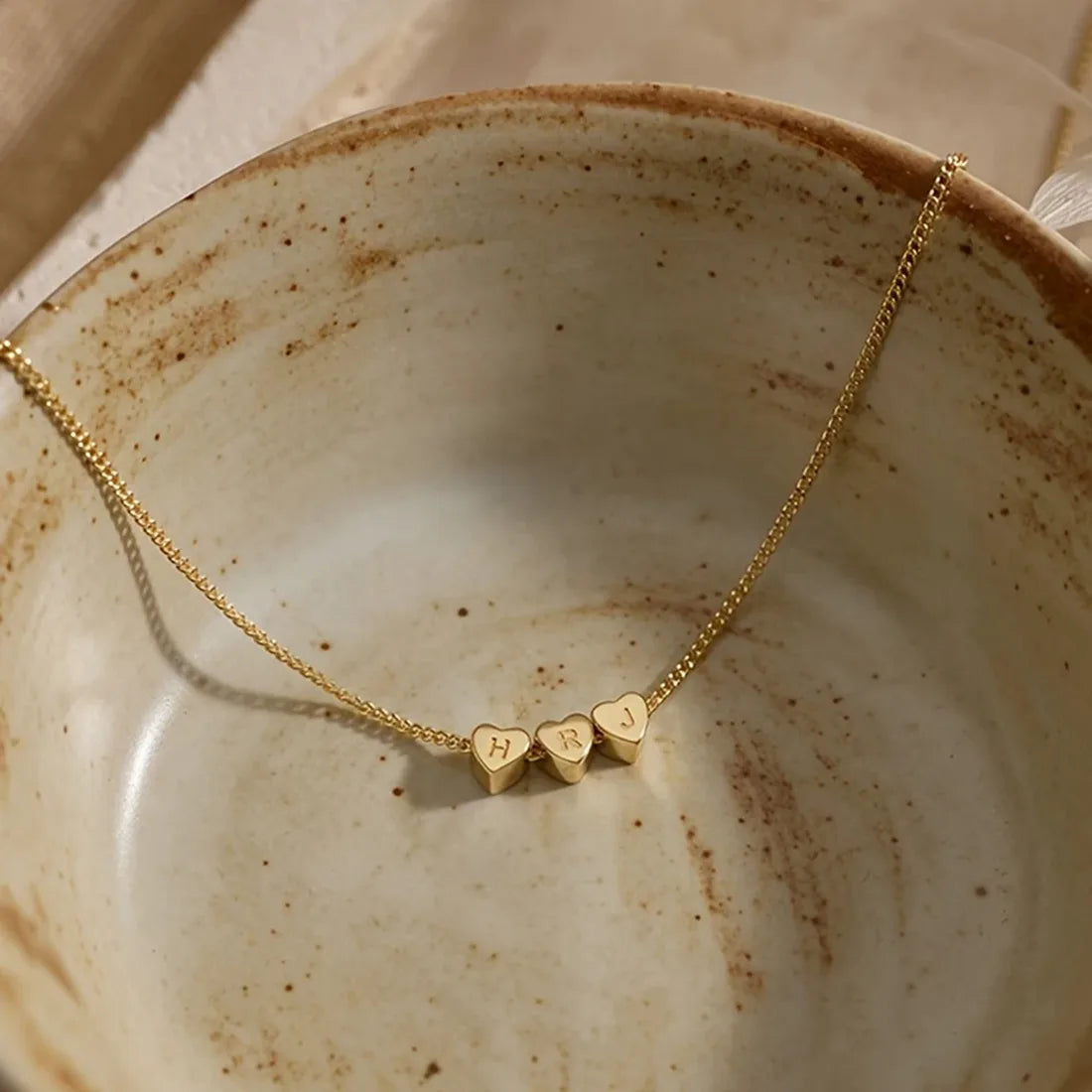 Fine gold steel necklace with two heart pendants engraved with initials, on a ceramic plate.